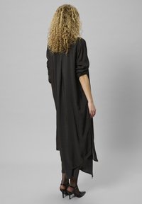 O femeie cu păr blonde ondulat, îmbrăcată cu o rochie lungă neagră peste pantaloni scurți și pantofi cu toc înalt negri, întoarsă cu fața.