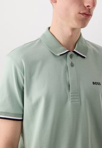 Světlězelená polo košile s texturovaným límcem, černobílými detaily a knoflíkovým zapínáním. Na prsou má logo.