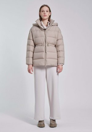 Vrouw die een beige gewatteerde jas met capuchon draagt, wijde witte broek en beige laarzen, staand tegen een effen lichte achtergrond.