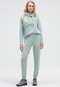 Hellgrüner weicher Stoff-Trainingsanzug mit Kordelzug-Kapuze, versehen mit einer aufgedruckten Botschaft. Jogginghosen mit elastischen Bündchen. Graue Sportschuhe.