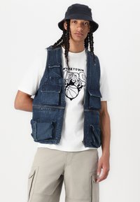 Denim darba veste ar vairākiem priekšējiem kabatām, uzvilkta virs baltas grafikas t-kreklu un kombinēta ar bēšiem cargo šortiem. Melns cepures aksesuārs.