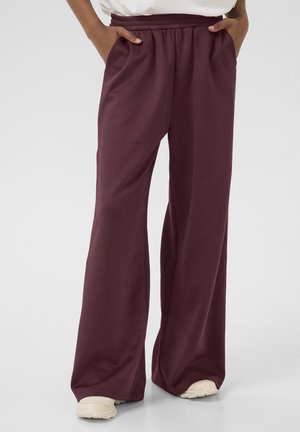 Pantalon bordeaux ample à taille haute avec une ceinture élastique, porté avec des chaussures blanches et les mains dans les poches sur un fond uni.