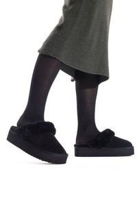 Pantofole nere foderate in pelliccia sintetica con suola a piattaforma, caratterizzate da una tomaia morbida e una fascia larga, abbinate a collant neri e a un vestito verde.