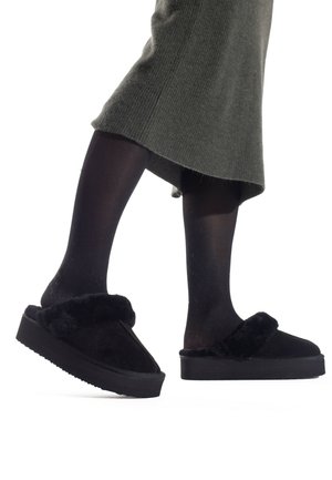 Pantofole nere foderate in pelliccia sintetica con suola a piattaforma, caratterizzate da una tomaia morbida e una fascia larga, abbinate a collant neri e a un vestito verde.