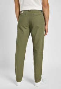 Oliwkowe spodnie o prostym kroju z dwiema tylnymi kieszeniami i metką na pasku. Noszone z białymi trampkami. Gładka struktura materiału.