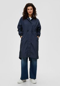 Granatowa wodoodporna kurtka z regulowanym paskiem w talii, dwoma przednimi kieszeniami i ściągaczami przy mankietach, zestawiona z szerokimi dżinsami i białymi trampkami.