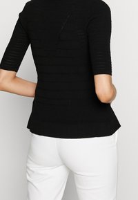 Černý pletený top s krátkými rukávy, který má žebrovanou texturu, otevřený zadní díl a peplum lem. V kombinaci s bílými slim kalhotami.