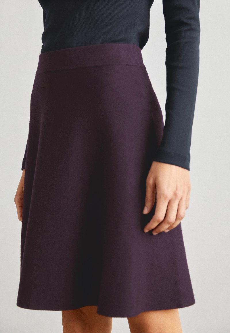 Femme portant une jupe trapèze violet foncé et un haut noir à manches longues, debout avec une main détendue le long du corps.