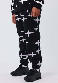 Joggers negros con un patrón de estrellas blancas, hechos de una tela suave. Cuentan con puños elásticos y un logo en el muslo. Combinados con zapatillas negras.