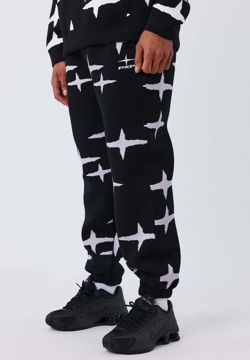 Joggers negros con un patrón de estrellas blancas, hechos de una tela suave. Cuentan con puños elásticos y un logo en el muslo. Combinados con zapatillas negras.