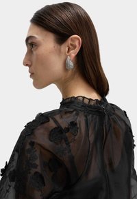 Duże kolczyki w kształcie łez, ozdobione błyszczącymi zdobieniami, zestawione z przezroczystą czarną koronkową bluzką z kwiatowymi wzorami i wysokim kołnierzem.