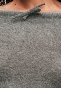 Prim-plan al unei persoane care poartă un pulover moale, pufos, gri, cu umerii lăsați și un mic fundiță legată la gât.