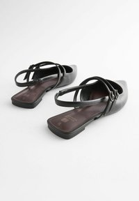Czarne płaskie buty z spiczastym noskiem, z otwartym tyłem i dwoma zaokrąglonymi paskami krzyżującymi się nad kostką. Gładkie wykończenie ze skóry.