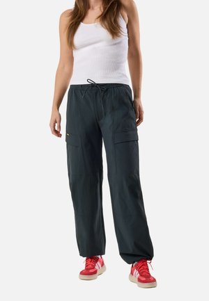 Kvinde iført hvid tanktop, sorte cargobukser med snøre i taljen og røde sneakers, stående mod en ensartet lys baggrund.