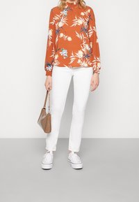 Blusa floreale a maniche lunghe arancione, con dettagli blu e bianchi, abbinata a jeans bianchi a gamba dritta e sneakers bianche. Borsa marrone.