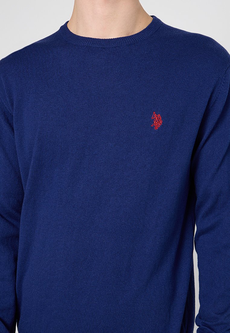 Dunkelblauer Strickpullover mit Rundhalsausschnitt und langen Ärmeln. Mit einem kleinen roten Logo auf der linken Brustseite. Weiche Textur.