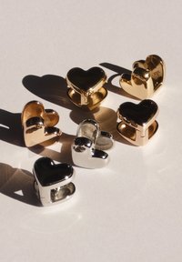 Seks hjerteformede charms i guld- og sølvnuancer, med polerede overflader og en metallisk glans, arrangeret på en lys baggrund.