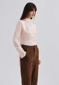 Light pink long-sleeve shirt with a high collar, made from smooth fabric with vertical stripes. Brown, loose-fitting pants with pockets.