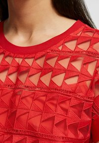 Top de malla roja con diseño de corte geométrico y detalles bordados texturizados. Presenta un escote redondo y mangas cortas.