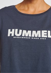 Námořnická modrá bavlněná tričko s širokým výstřihem, zdobené velkým bílým logem "HUMMEL" a sloganem "SPORTOVNÍ MÓDA OD ROKU 1923."