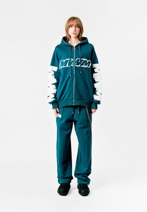Sudadera con cremallera en color azul verdoso con texto gráfico blanco, puños elásticos. Combinada con pantalones de corte holgado a juego, que cuentan con un bolsillo lateral y un acento de cadena.