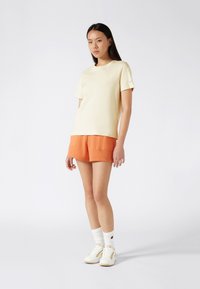 T-shirt jaune clair à manches courtes associé à un short orange, avec une texture lisse et un design minimal. Baskets blanches avec des accents dorés.
