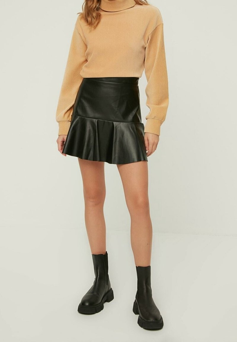 Jupe évasée en simili cuir noir avec un ourlet froncé, associée à un pull en maille beige à côtes et des bottes noires jusqu'aux genoux.