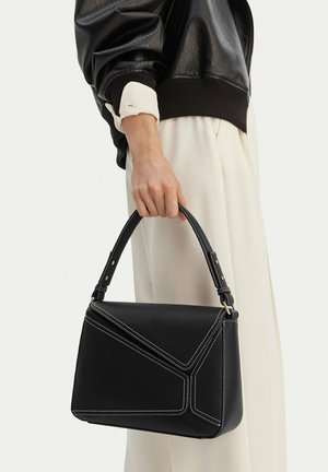 Donna con giacca di pelle nera e pantaloni bianchi tiene in mano una borsa nera texturizzata con cuciture bianche e manico corto.