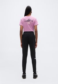 Άτομο που στέκεται με την πλάτη προς τα εμπρός, φορώντας ροζ μπλουζάκι με σχέδιο, μαύρο αθλητικό παντελόνι και μαύρα αθλητικά παπούτσια, σε απλό λευκό φόντο.