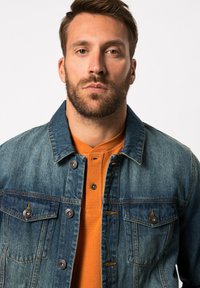 Chaqueta de mezclilla en azul deslavado con cierres de botón y dos bolsillos en el pecho, superpuesta a una camisa de color naranja con cuello que presenta una tela texturizada.