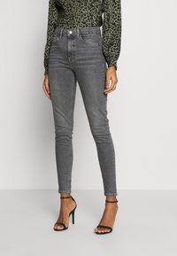 Jeans ajustados de tiro alto en denim gris oscuro. Presentan un diseño clásico de cinco bolsillos y un ligero desgaste. Combinados con tacones negros con tiras.