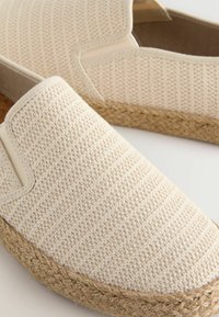 Beige espadriller med struktureret stofoverdel, elastiske sidedele og flettede jutesåler. Simpel, slip-on design. Flad profil.