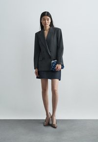 Blazer gris à rayures fines croisé, associé à une jupe en denim foncé. Talons pointus marron et une petite pochette bleue à motifs.
