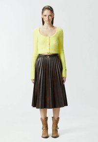 Žuta pahuljasta skraćena kardigan s okruglim izrezom, uparen s naboranom tamnosmeđom kožnom suknjom i svijetlosmeđim kaubojskim čizmama.