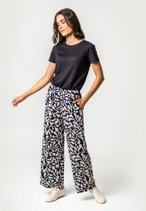 Woman wearing plain black short-sleeve shirt, wide-leg pants with abstract black and white pattern, and white sneakers, standing with hand in pocket.