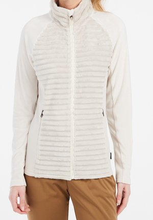 Femme portant une veste polaire crème à fermeture éclair intégrale, avec une texture côtelée horizontale et des poches zippées, associée à un pantalon marron.