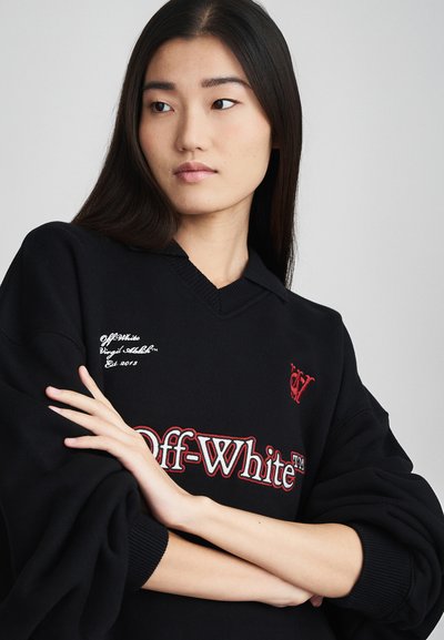 Zwarte oversized trui met een V-halsontwerp. Beschikt over witte en rode geborduurde logo's en tekst op de borst en mouw. Zachte textuur.
