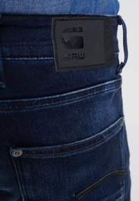 Tumši zili džinsi ar redzamu aizmugures kabatu, jostu cilpu un melnu ādas zīmolu, uz kura ir iepresēts "G RAW" logo.