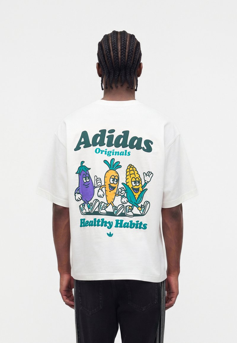 Person mit geflochtenem Haar, die ein weißes Adidas-T-Shirt mit Cartoon-Aubergine, Karotte und Mais trägt, mit dem Text „Gesunde Gewohnheiten“ auf dem Rücken.