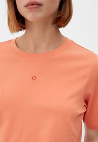 Kvinde iført en ferskenfarvet T-shirt med et lille, abstrakt broderet design, der viser to sammenkoblede cirkler på brystet.