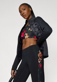 Casaco atlético preto sobre um top de desporto floral e leggings de cintura alta. As leggings apresentam um padrão floral colorido na lateral.