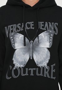 Hanorac negru cu un grafic mare de fluture gri centrat pe față, cu textul "VERSACE JEANS COUTURE" deasupra și dedesubt, tot în gri.