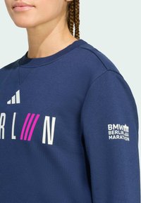 Felpa blu navy con scollo a girocollo, caratterizzata da testo bianco "BERLIN" e strisce rosa. La manica sinistra presenta il logo bianco "BMW Berlin Marathon".