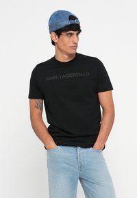 Črna bombažna majica s kratkimi rokavi in okroglim izrezom, na katerem je tonirana oznaka "KARL LAGERFELD" na prsih. Nosi se z modrim jeans kapo.