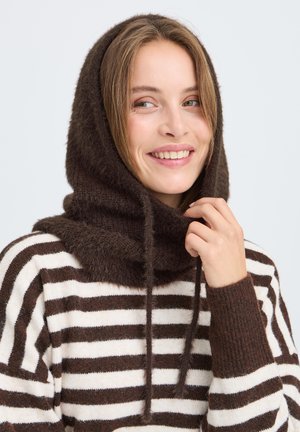 Jeune femme souriante, portant une écharpe marron duveteuse à capuche et un pull rayé blanc et marron, tenant le cordon de l'écharpe.