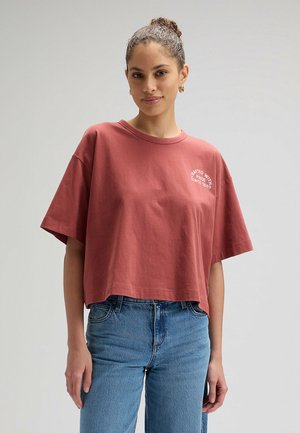 Vrouw met haar in een knot, draagt een losse, korte t-shirt in roestkleur en een hoge taille blauwe jeans, staand tegen een effen grijze achtergrond.