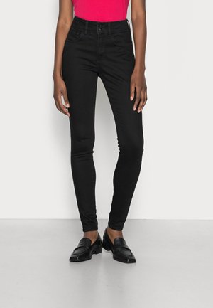 Kobieta ubrana w czarne, obcisłe, wysokie spodnie jeansowe, czarne mokasyny i różową bluzkę, stojąca na jednolitym jasnym tle.