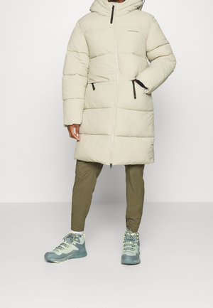 Osoba ubrana w długi beżowy pikowany płaszcz z kapturem, oliwkowozielone spodnie i zielone buty trekkingowe, stojąca na tle jednolitego jasnego tła.