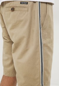 Shorts beiges en coton, avec une poche arrière à bouton et une bande latérale en noir et blanc. Texture lisse et design ajusté.