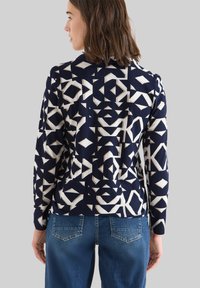 Chaqueta de color azul marino con patrón de formas geométricas en color crema, con mangas largas y un ajuste ligeramente entallado, combinada con pantalones vaqueros de color azul claro.